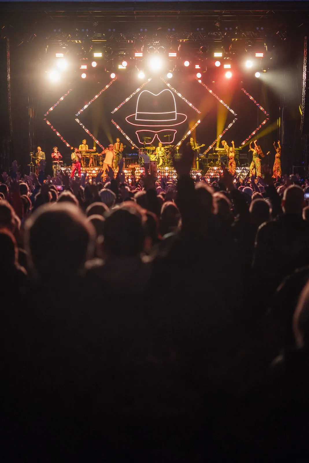 Publikum mit erhobenen Händen vor der hell beleuchteten Konzertbühne mit Band und großem Hut-Brillen-Symbol.
