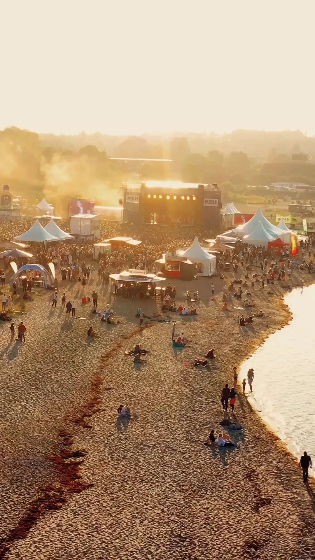 Luftaufnahme eines Strandkonzerts mit Bühne, Zelten und dichtem Publikum im goldenen Abendlicht am Wasser.
