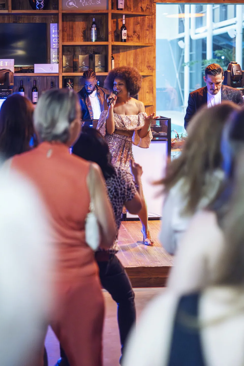 Sängerin im glitzernden Kleid mit Mikrofon vor Gästen und Musikern in einer Bar mit warmem Licht.