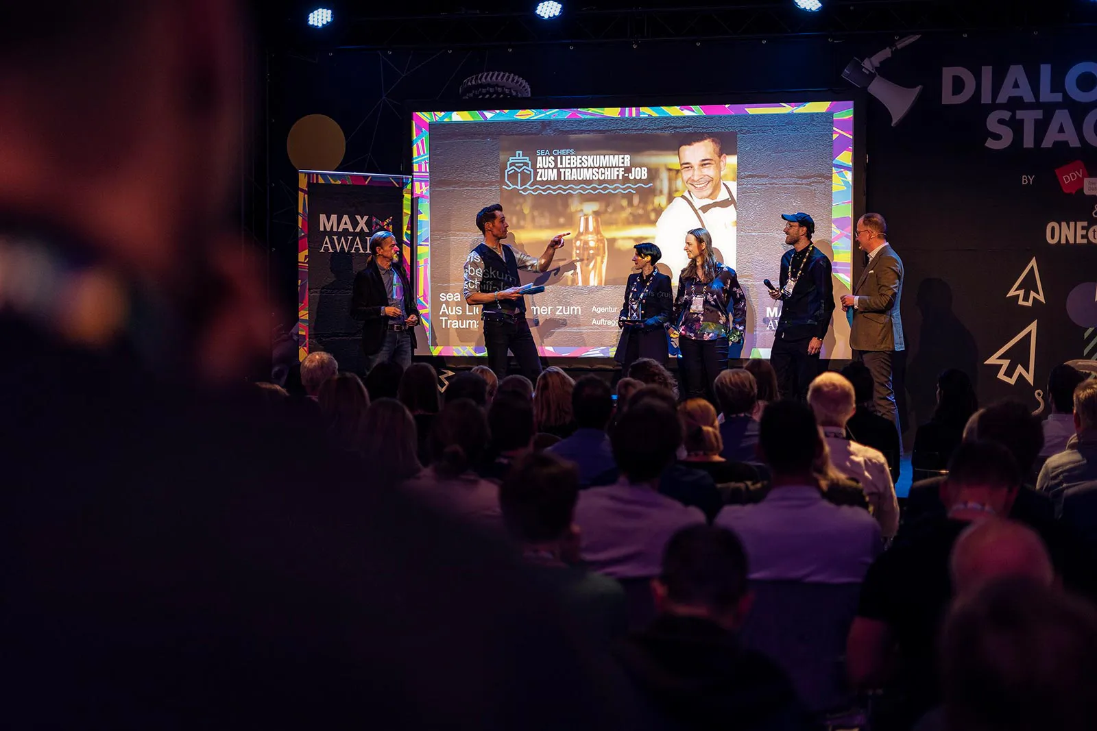 Sechs Personen auf der beleuchteten Bühne beim MAX Award, davor sitzendes Publikum, dahinter eine große Leinwand.