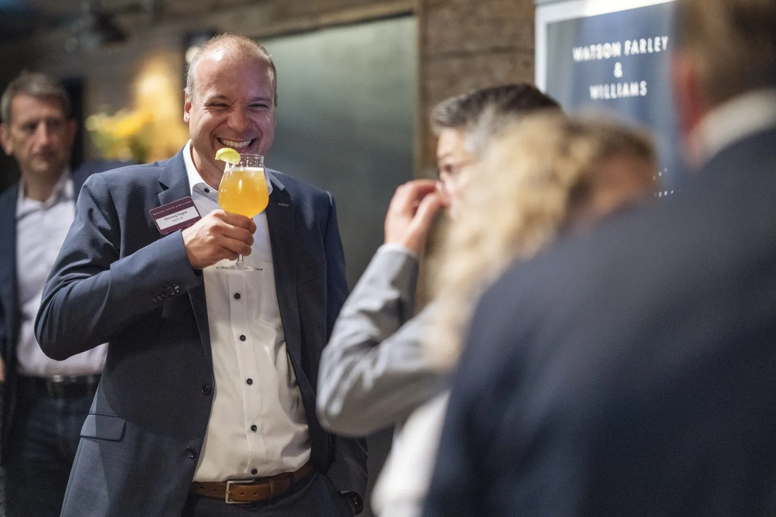 Lachender Mann im Anzug hält ein Glas mit orangem Getränk, umringt von Gästen bei einem Event in warmem Licht.