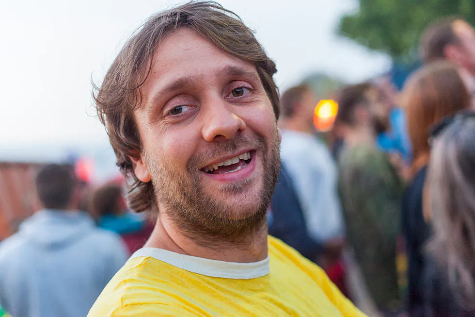 Lächelnder Mann mit Bart im gelben T-Shirt vor unscharfem Festivalpublikum im Freien.