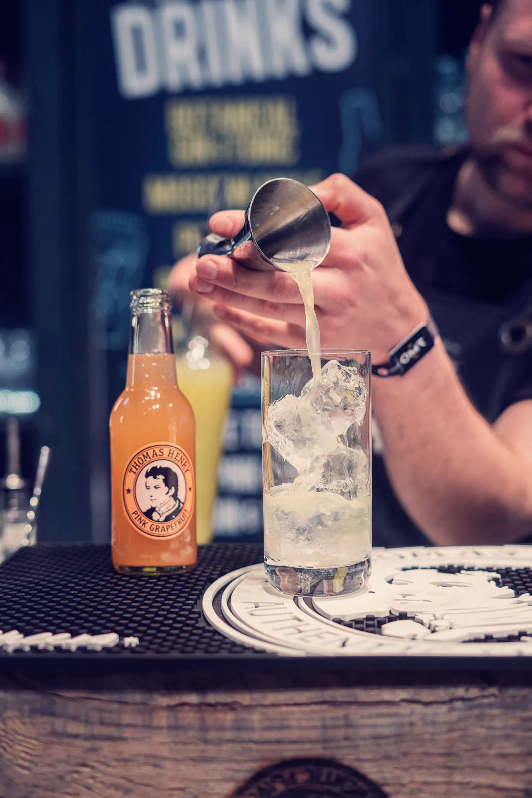 Barkeeper gießt einen Drink in ein Glas mit Eis; daneben steht Thomas Henry Pink Grapefruit auf der dunklen Theke.