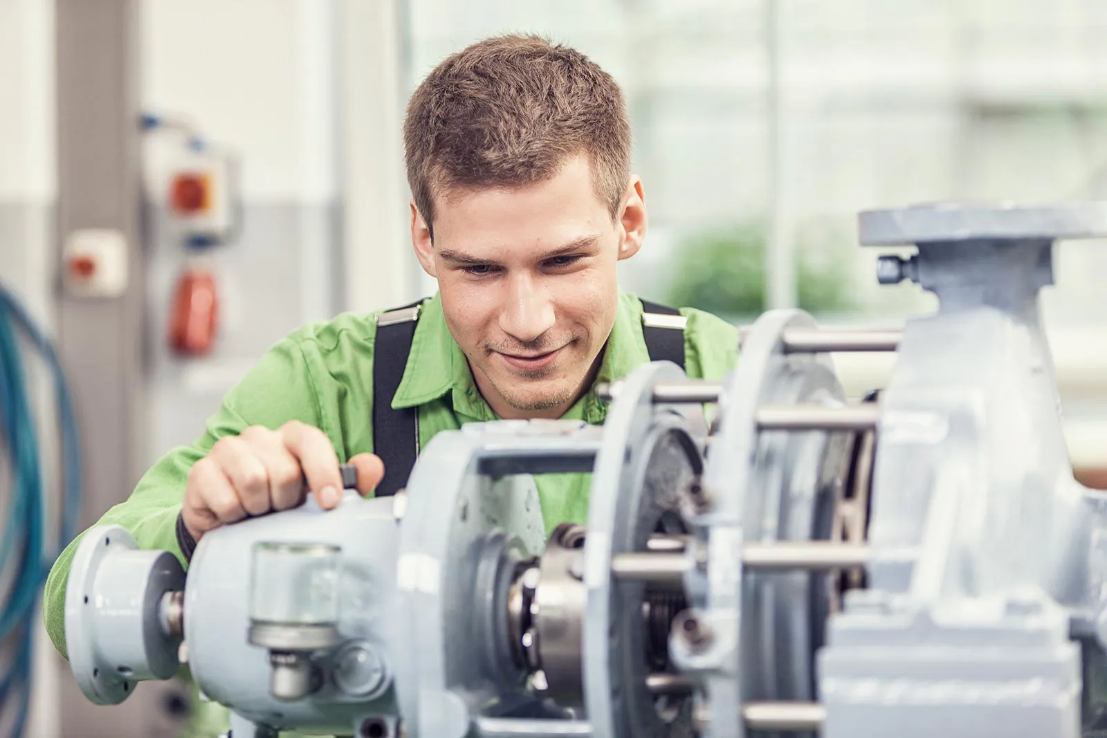 Junger Mann in grüner Arbeitskleidung prüft eine geöffnete Industriepumpe in einer hellen Werkstatt.