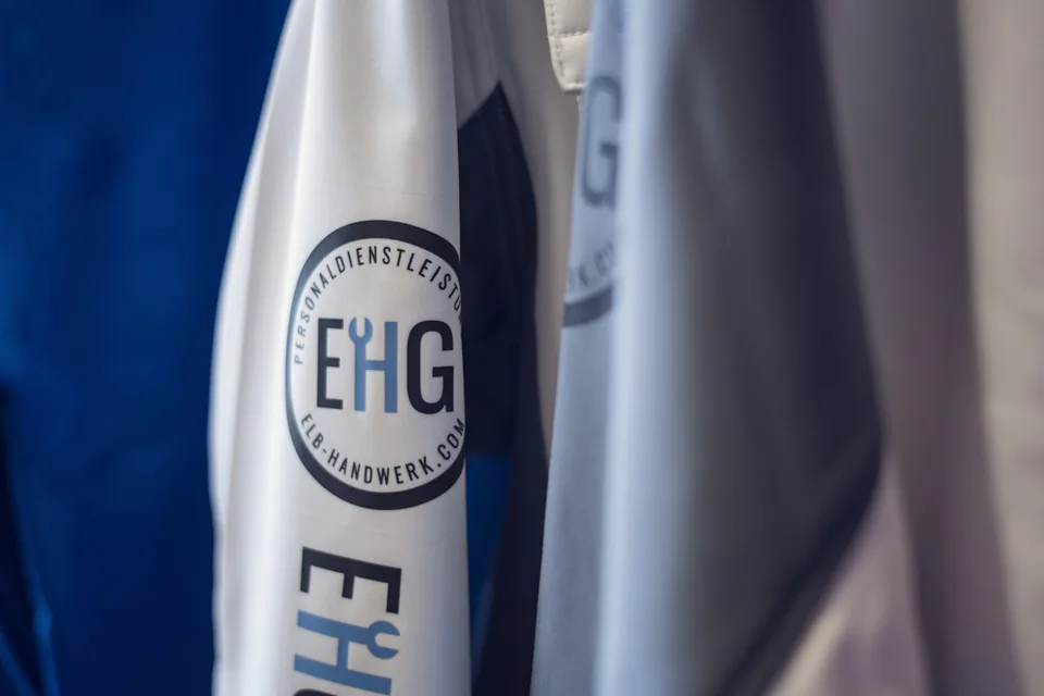 Nahaufnahme hängender weißer und grauer Arbeitskleidung mit EHG-Logo von Elb Handwerk in weichem Seitenlicht.