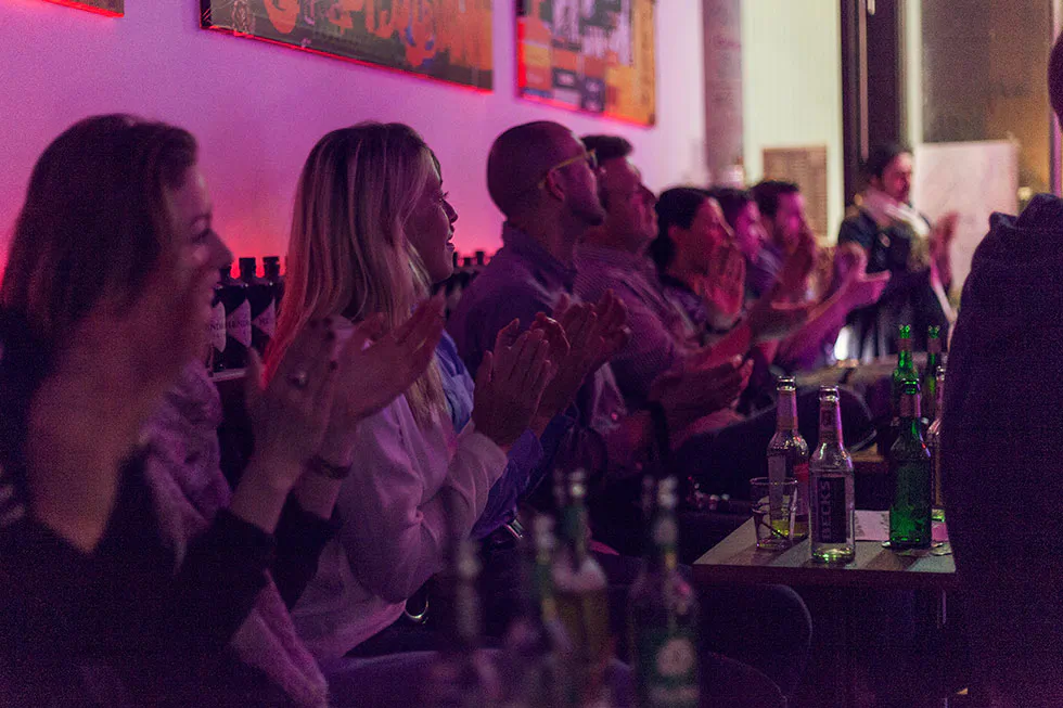 Publikum applaudiert an Tischen mit Getränkeflaschen in einem kleinen, pink beleuchteten Veranstaltungsraum.