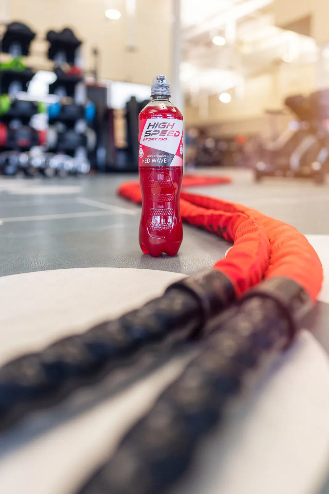 Rote Sportgetränkflasche neben orangefarbenen Trainingsseilen auf dem Boden eines hellen Fitnessstudios.