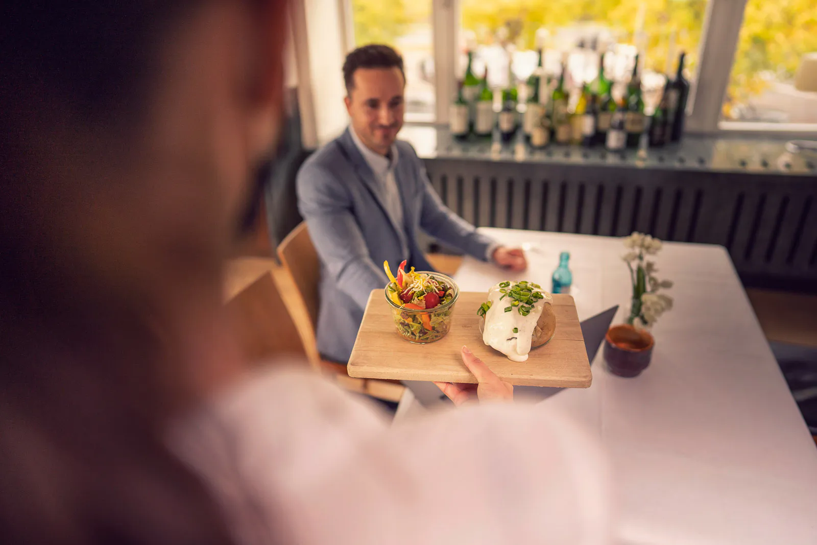 Servicekraft bringt Salat und Ofenkartoffel auf einem Holzbrett zu einem Gast am Tisch im hellen Restaurant.
