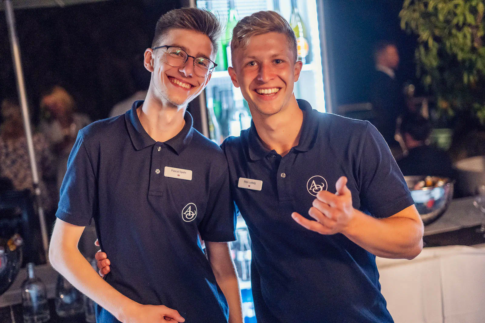 Zwei lächelnde Männer in dunkelblauen Poloshirts an einer Bar vor beleuchtetem Kühlschrank; einer zeigt eine Handgeste.