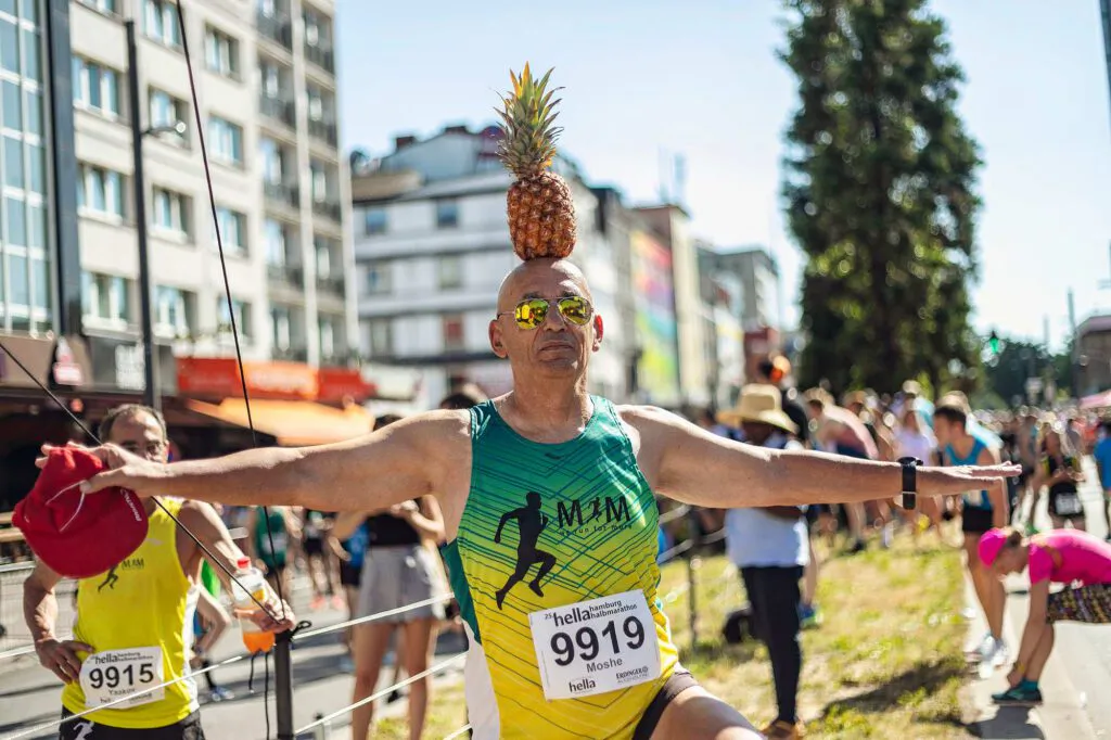 Läufer mit Startnummer 9919 balanciert eine Ananas auf dem Kopf und breitet auf sonniger Stadtstraße die Arme aus.