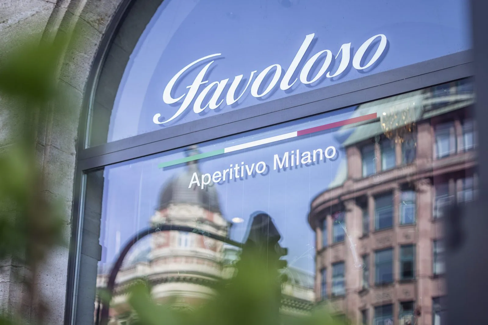 Rundbogenfenster mit Schriftzug Favoloso Aperitivo Milano, gespiegelten Gebäuden und unscharfen Blättern im Vordergrund.