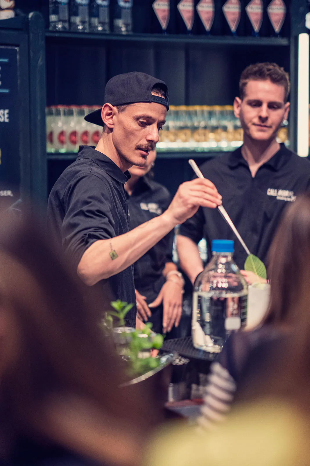 Barkeeper mit schwarzer Kappe bereitet einen Drink am Tresen zu, dahinter Kollegen und beleuchtete Flaschenregale.