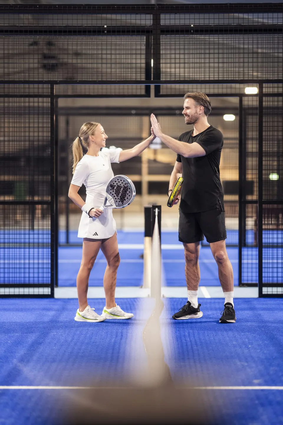 Frau und Mann mit Padelschlägern klatschen sich am Netz auf einem blauen Indoor-Padelplatz ab.