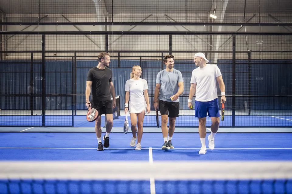 Vier Personen mit Padelschlägern gehen lächelnd über einen blauen Indoor-Padelplatz unter Hallenlicht.