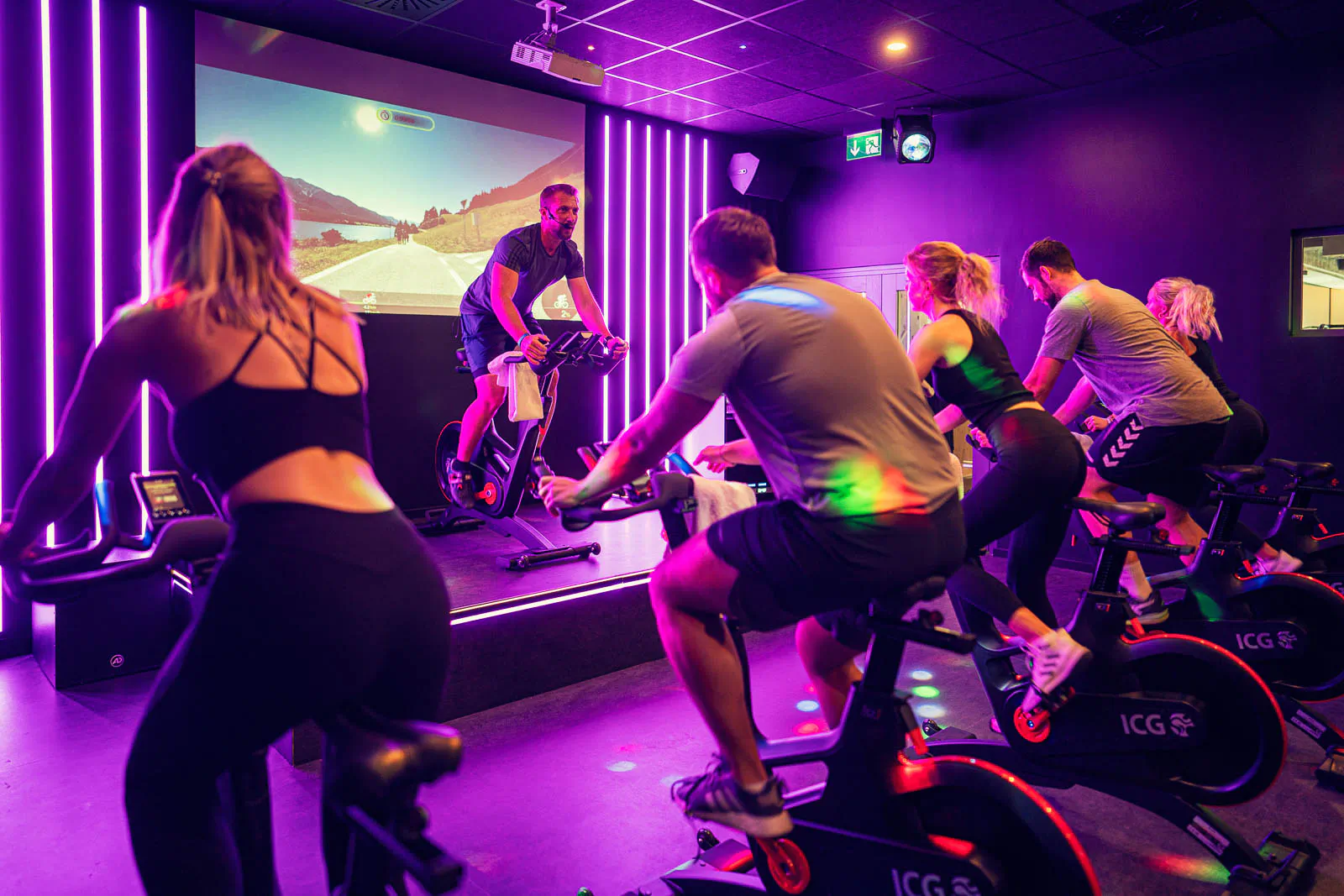 Gruppe trainiert auf Indoor-Bikes im violett beleuchteten Fitnessraum, Trainer auf Podest vor einer Leinwand.