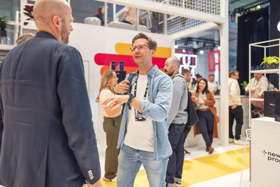 Zwei Männer im Gespräch am hell beleuchteten Awin-Messestand, weitere Besucher im Hintergrund.