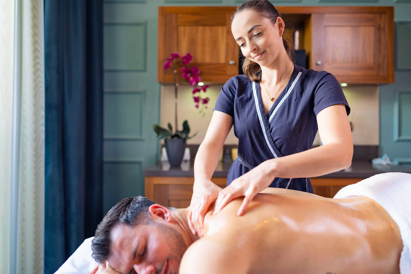 Frau massiert den eingeölten Rücken eines Mannes auf einer Massageliege im seitlichen Fensterlicht.