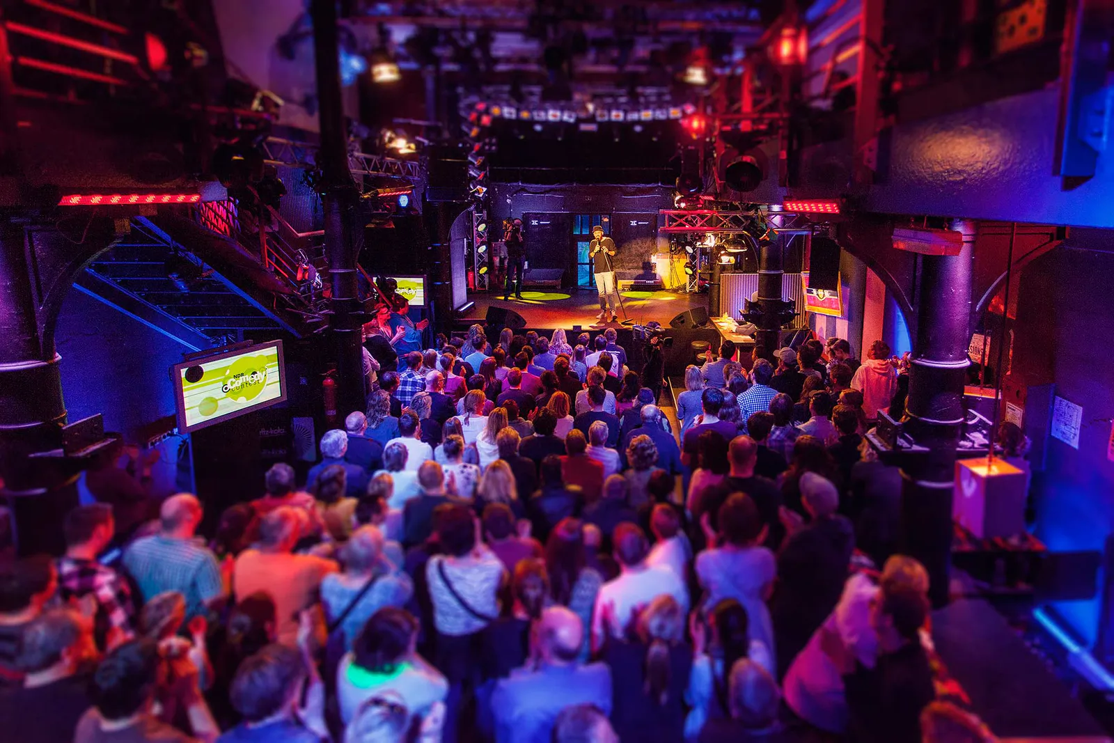 Publikum vor der Bühne beim NDR Comedy Contest, ein Comedian im Scheinwerferlicht, der Saal rot und blau beleuchtet.