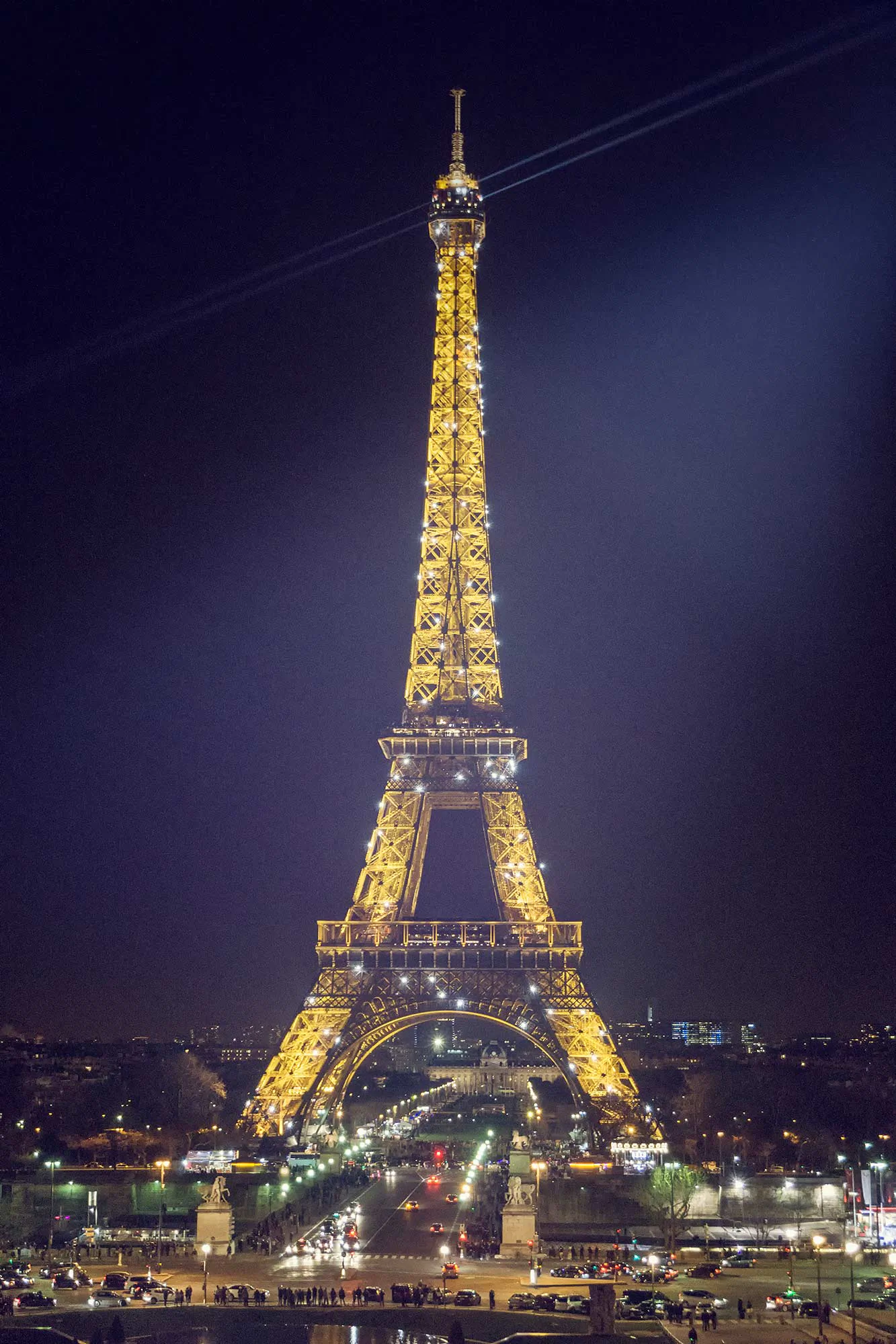 Goldgelb beleuchteter Eiffelturm bei Nacht, davor Straßenverkehr und kleine Menschengruppen.
