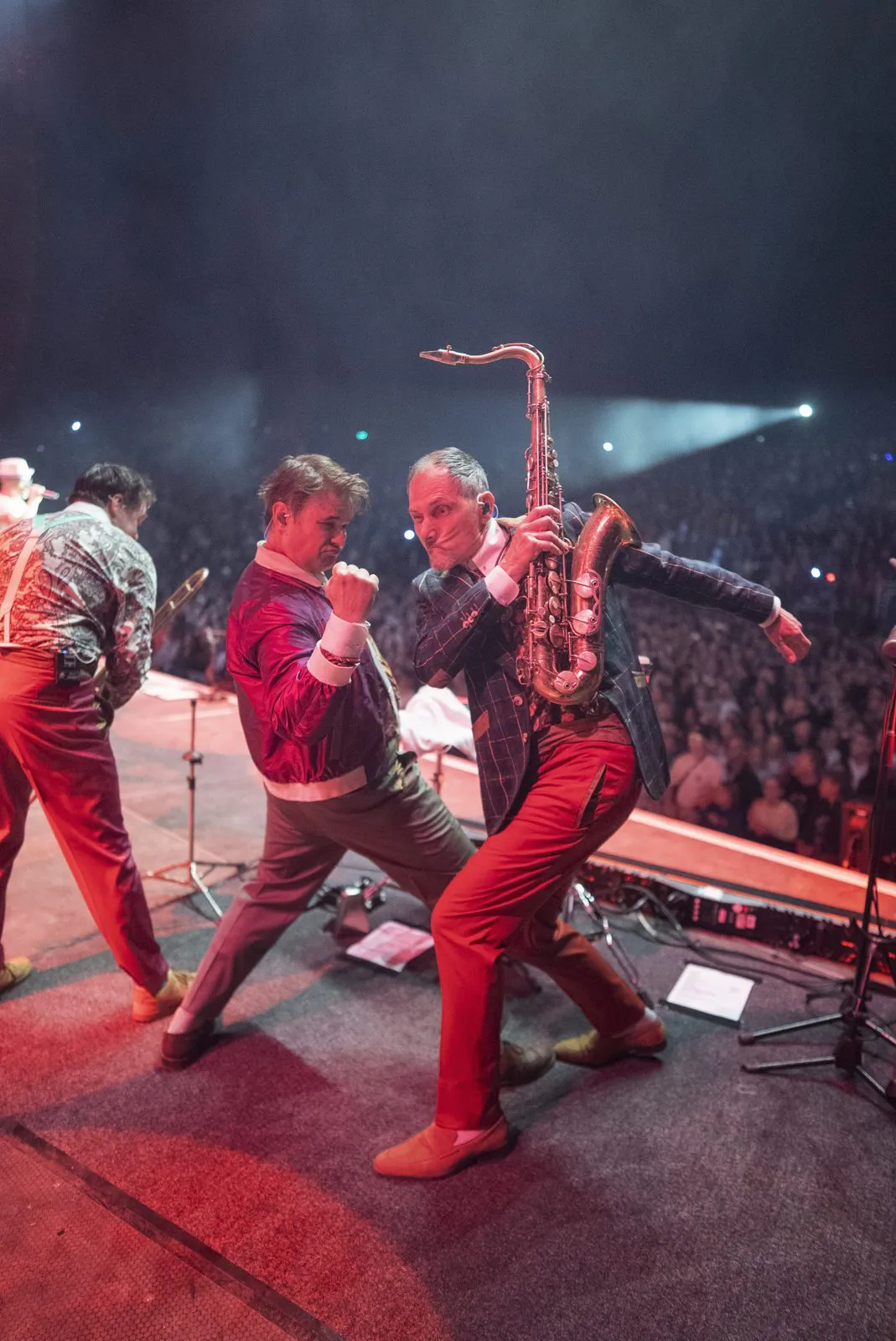 Musiker posieren neben einem Saxofonisten auf der rot beleuchteten Konzertbühne vor dichtem Publikum.