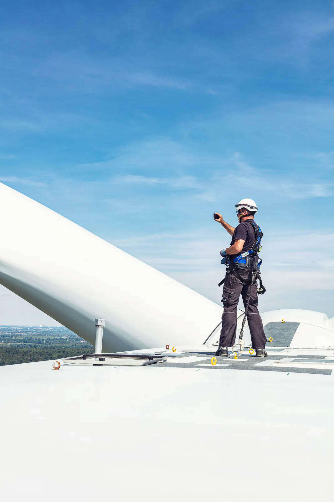 Techniker mit Helm und Sicherungsgurt steht bei Tageslicht auf einer Windkraftanlage neben einem Rotorblatt.