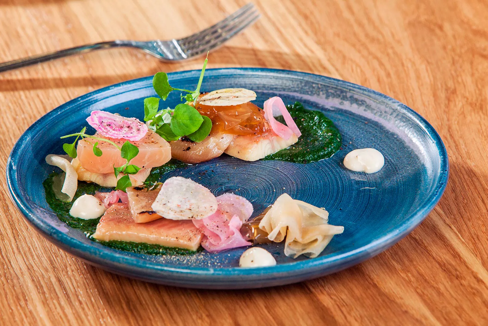 Blauer Keramikteller mit angerichtetem Fisch, Kräutern, eingelegtem Gemüse und Saucen auf einem Holztisch.