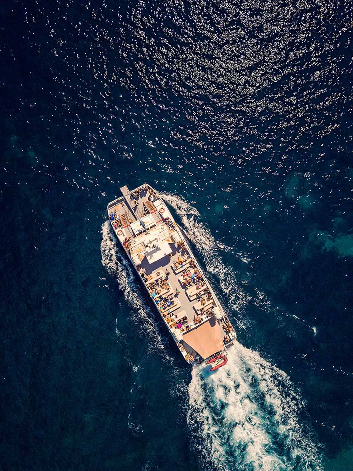 Drohnenaufnahme eines Boots mit Menschen an Deck und weißer Kielwasserspur auf dunkelblauem Wasser im Sonnenlicht.