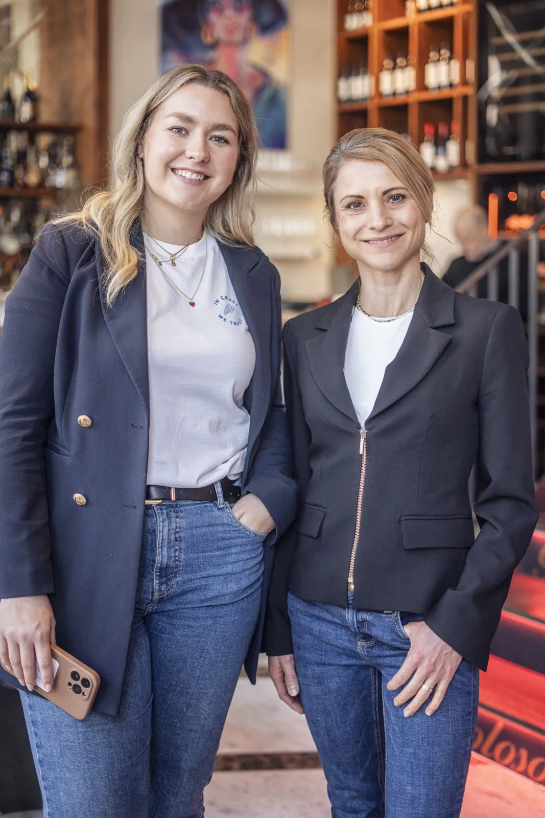 Zwei lächelnde Frauen in dunklen Blazern und Jeans stehen in einem Restaurant vor Regalen mit Flaschen.