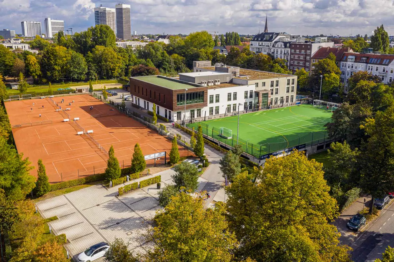 Drohnenansicht der Aspria-Sportanlage mit roten Tennisplätzen, grünem Fußballfeld und Bäumen im Sonnenlicht.