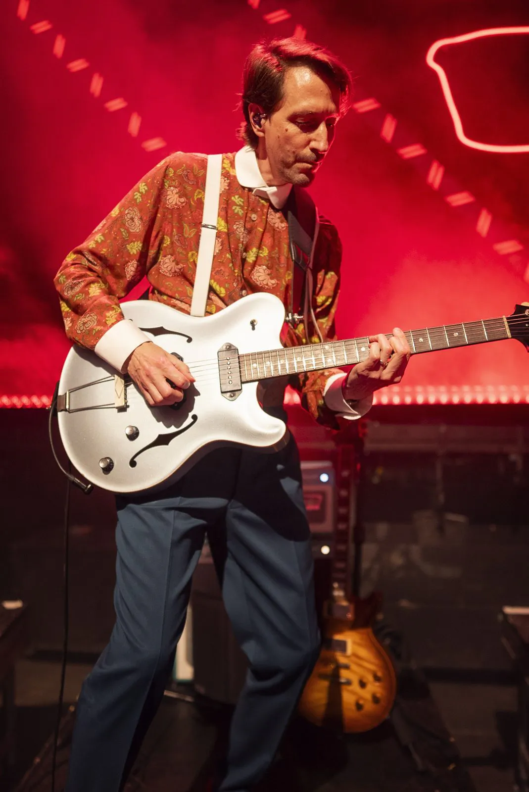 Gitarrist mit gemustertem Hemd und Hosenträgern spielt eine weiße E-Gitarre auf rot beleuchteter Konzertbühne.