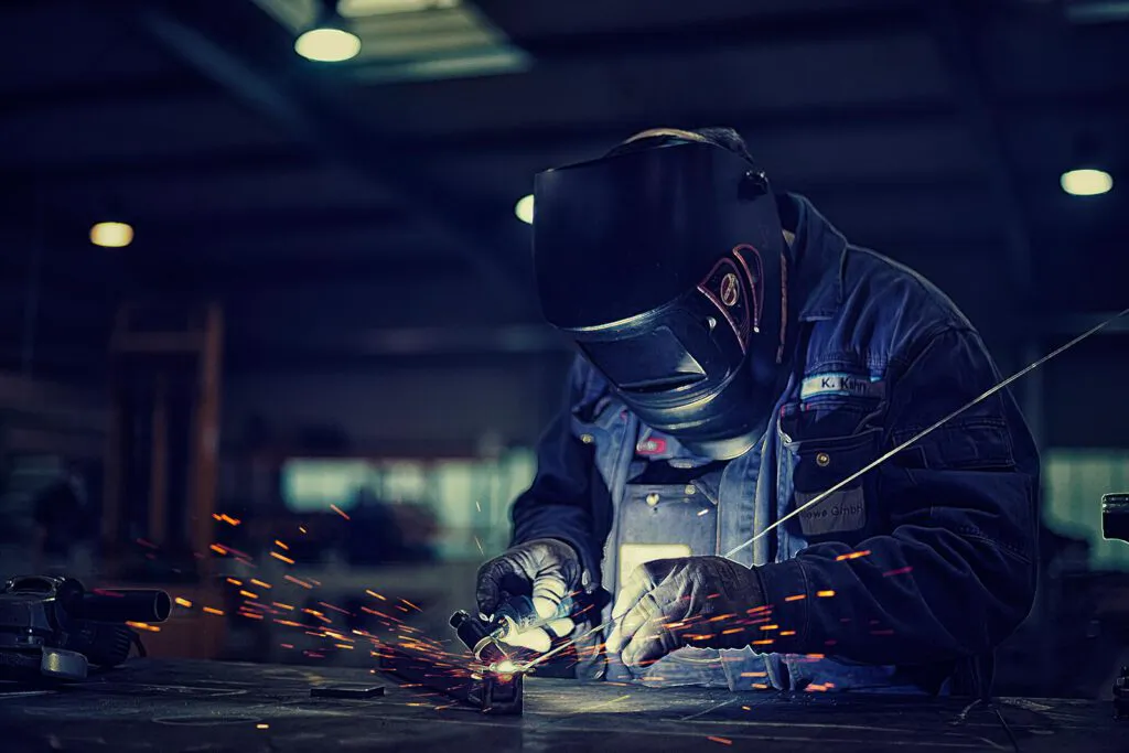 Schweißer mit Schutzhelm arbeitet bei Funkenflug an einem Metallstück in einer dunkel beleuchteten Werkhalle.