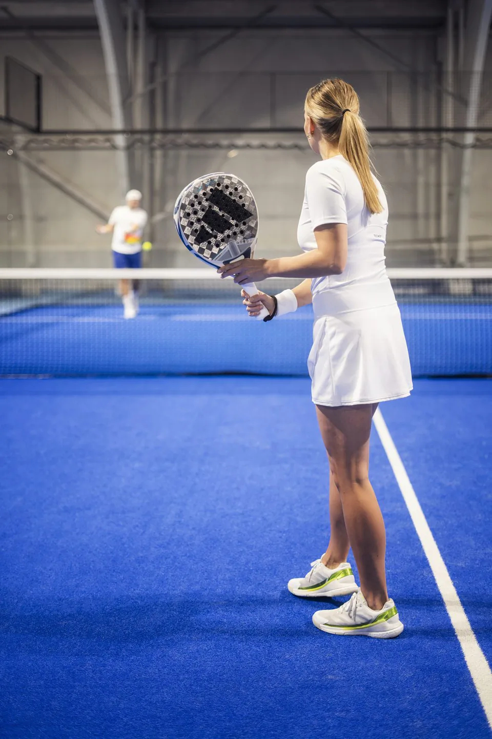 Frau in weißem Sportoutfit mit Padelschläger auf blauem Hallenplatz, gegenüber ein Spieler hinter dem Netz.