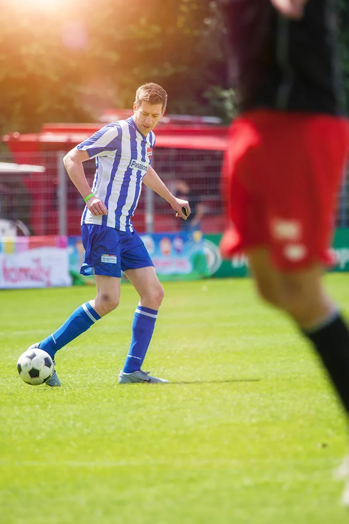 Fußballspieler im blau-weiß gestreiften Trikot am Ball auf sonnigem Rasen, unscharfer Spieler in Rot im Vordergrund.