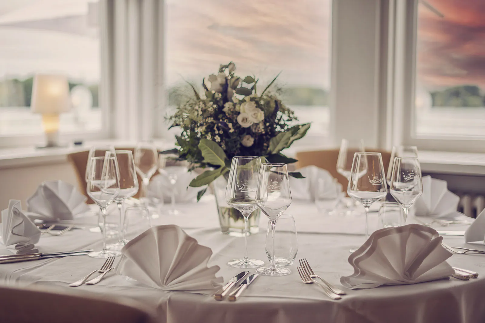 Festlich gedeckter Tisch mit Weingläsern, gefalteten Servietten und Blumenstrauß im hellen Gastraum.