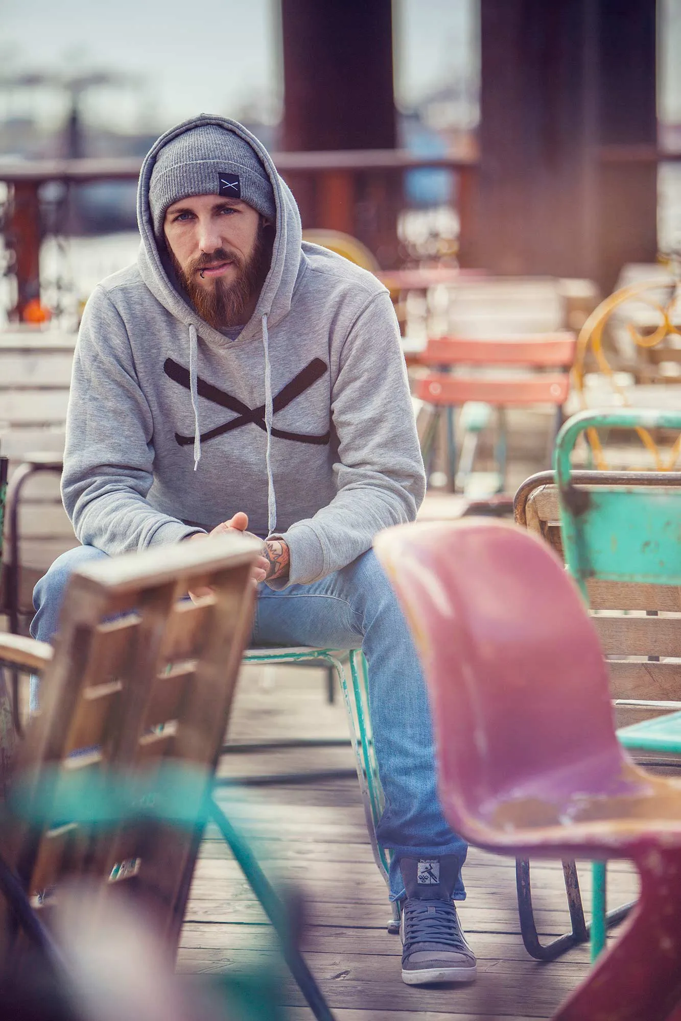 Ein bärtiger Mann mit grauem Hoodie und Mütze sitzt bei Tageslicht zwischen bunten Stühlen auf einer Holzterrasse.