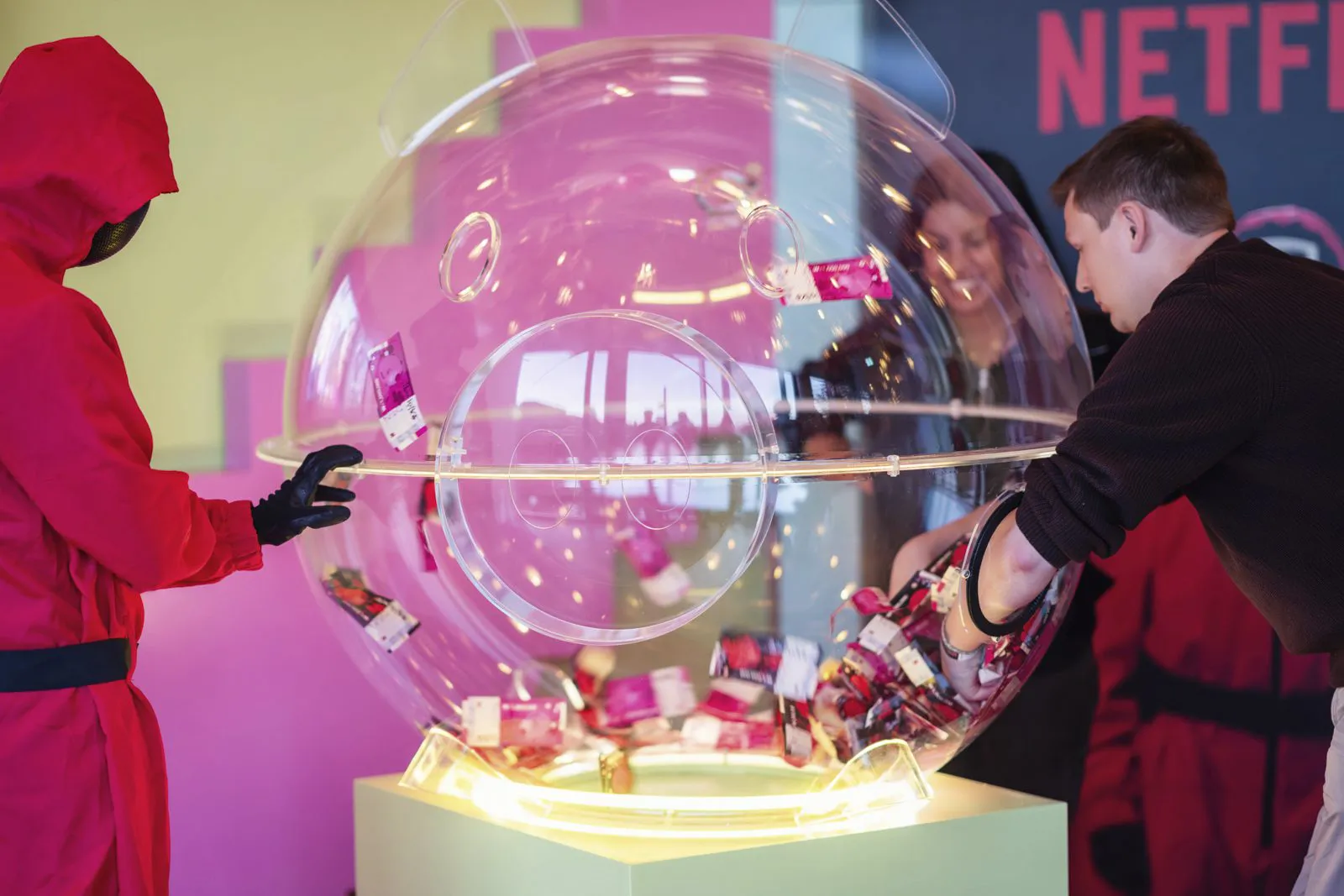 Ein Mann greift in eine beleuchtete Kugel mit Produkten, daneben eine rot maskierte Person beim Netflix-Event.