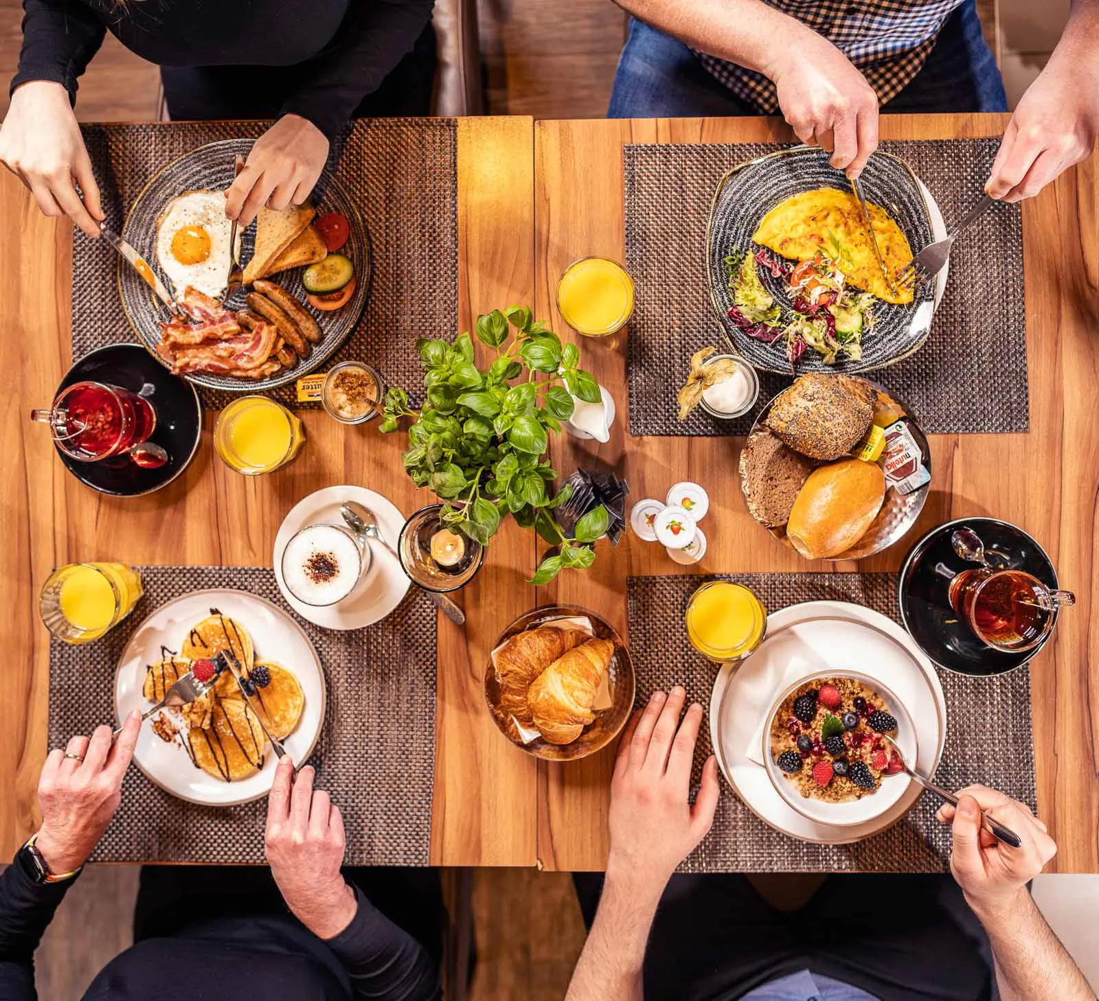 Vier Personen frühstücken am Holztisch mit Spiegelei, Speck, Omelett, Pancakes, Müsli, Brötchen und Orangensaft.