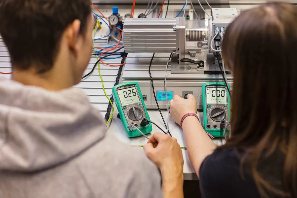 Zwei Personen bedienen einen elektrischen Versuchsaufbau mit Motor, Kabeln und zwei Multimetern im Technikraum.