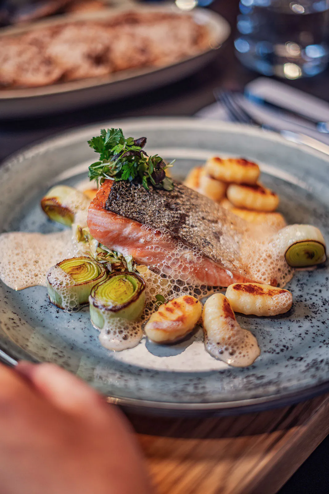 Gebratenes Lachsfilet mit knuspriger Haut, Lauch, Gnocchi und Schaumsauce auf einem grauen Teller im Restaurant.