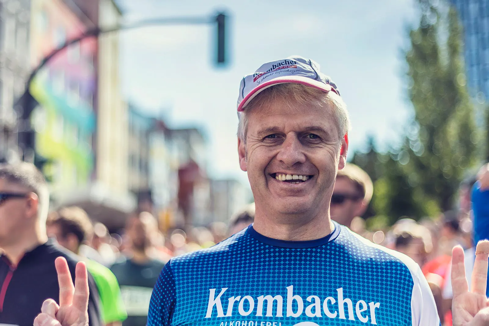Lächelnder Läufer mit Kappe und blauem Sportshirt zeigt im Sonnenlicht auf einer Straße beide Hände im Victory-Zeichen.