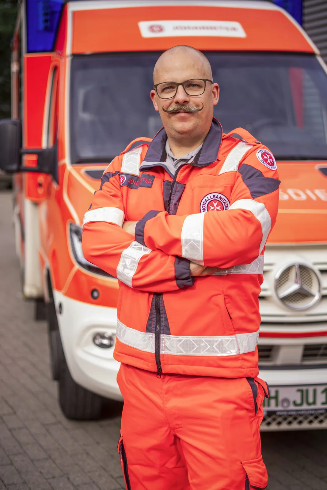Johanniter-Mitarbeiter mit Brille und Schnurrbart steht mit verschränkten Armen in Einsatzkleidung vor einem Rettungswagen.