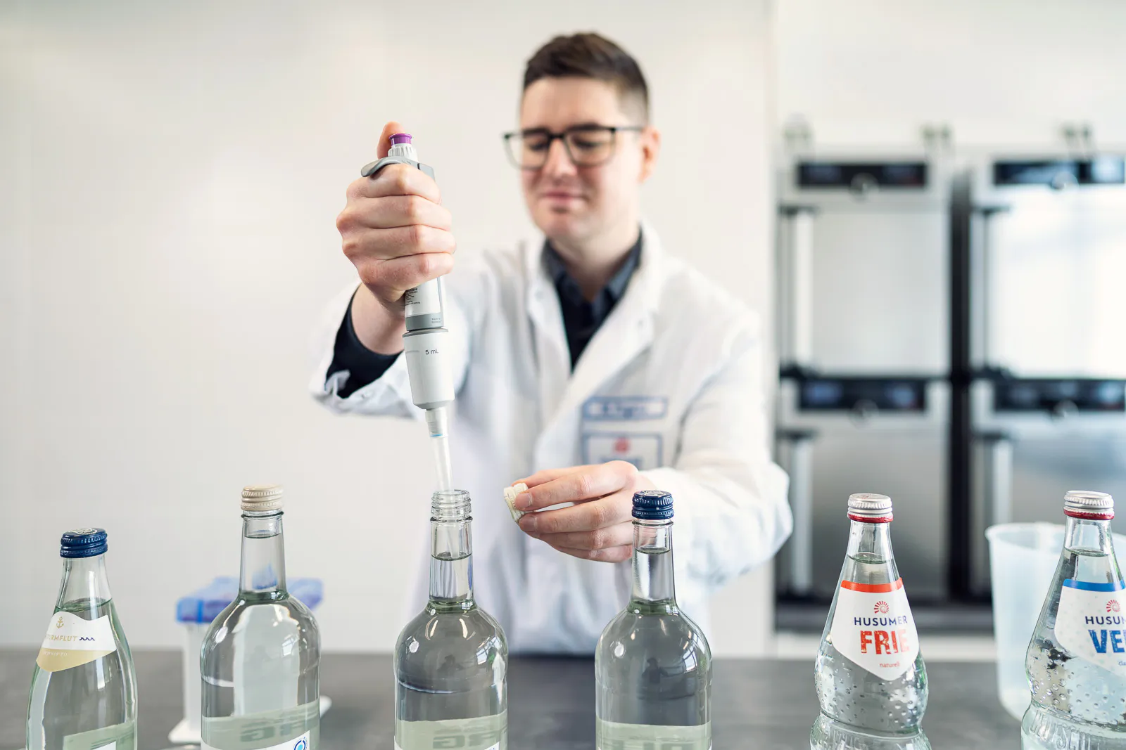 Mann im weißen Laborkittel pipettiert Flüssigkeit aus einer Glasflasche im hellen Labor des Husumer Mineralbrunnens.