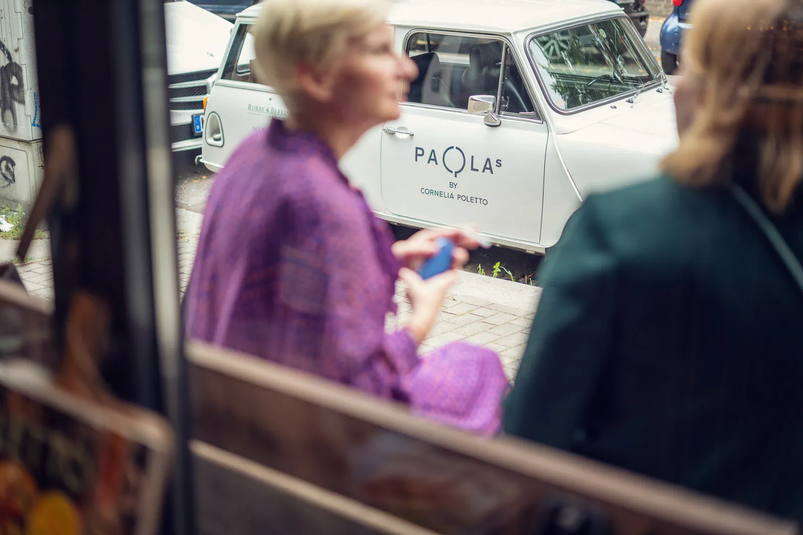 Zwei unscharfe Personen vor einem weißen Oldtimer mit der Aufschrift „PAOLA’S by Cornelia Poletto“ im Tageslicht.