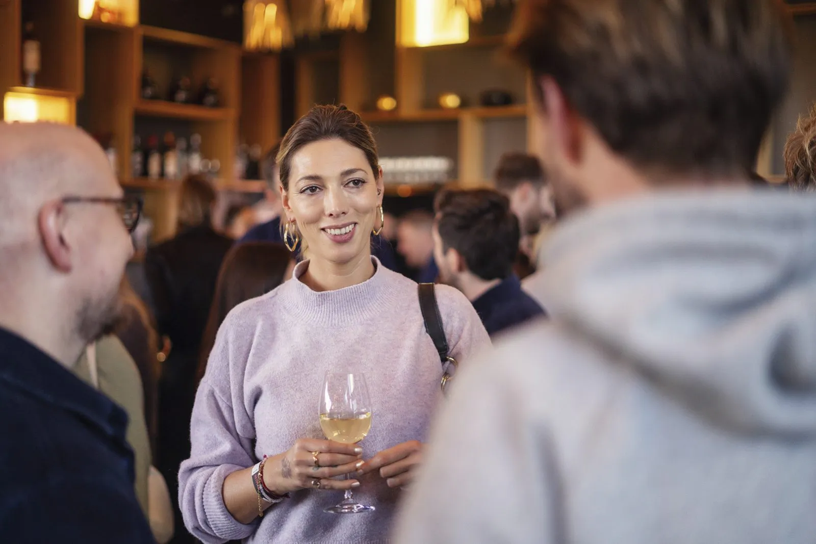 Frau im fliederfarbenen Pullover hält ein Weinglas und spricht mit Gästen in einer warm beleuchteten Bar.