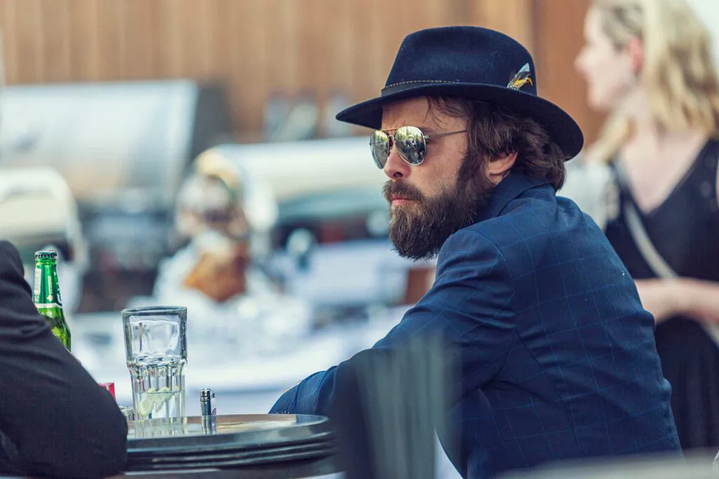 Bärtiger Mann mit dunklem Hut, Sonnenbrille und kariertem Sakko sitzt an einer Bar mit Gläsern und Flasche.