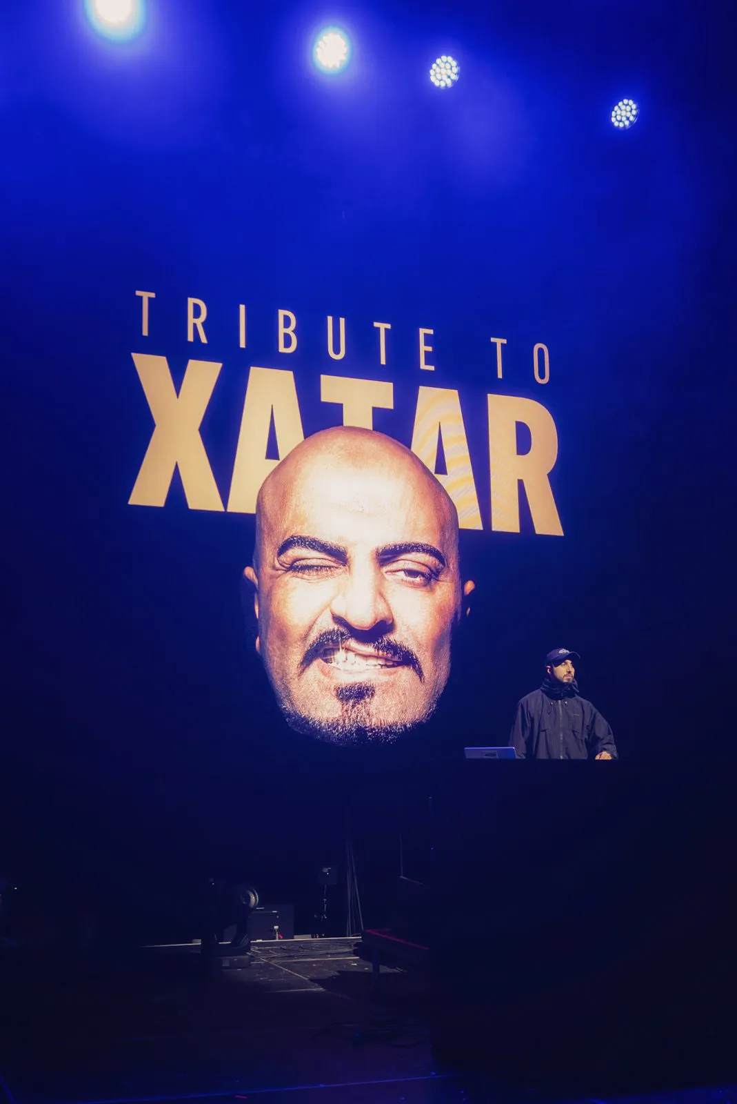 Blau beleuchtete Konzertbühne mit DJ vor großem Xatar-Porträt und dem Schriftzug Tribute to Xatar.