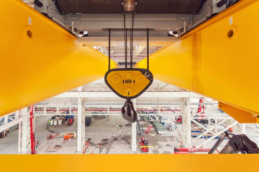 100-Tonnen-Kranhaken zwischen gelben Stahlträgern über einer hellen Industriehalle mit Personen und Arbeitsbühnen.