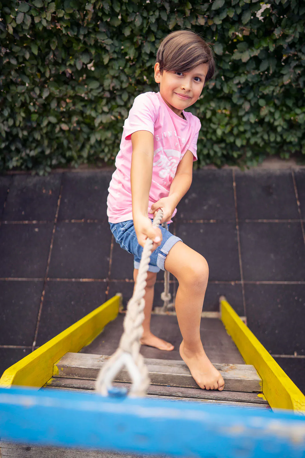 Barfüßiges Kind in rosa T-Shirt und Jeansshorts hält ein Seil auf einer bunten Spielplatzbrücke vor einer Hecke.
