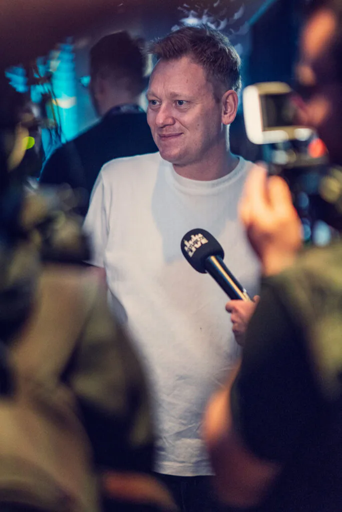 Mann im weißen T-Shirt gibt ein Interview vor Kamera und Mikrofon im blau beleuchteten Eventbereich.