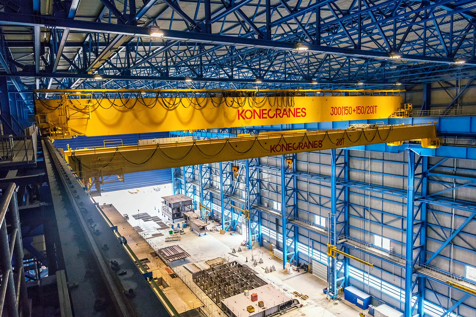Gelbe KONECRANES-Brückenkrane unter blauem Stahltragwerk in einer hell beleuchteten Industriehalle.